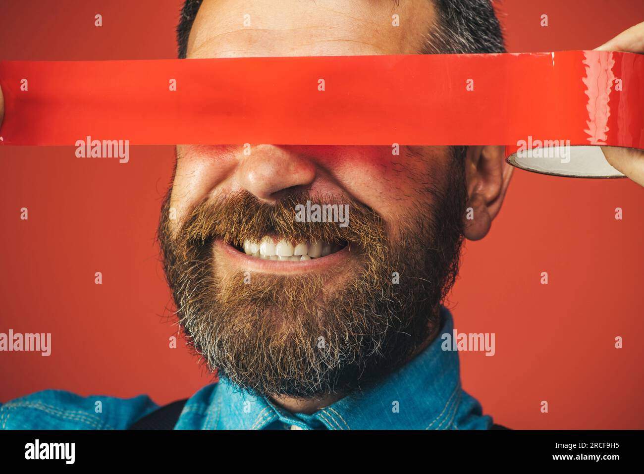 Smiling bearded man wrapping insulating tape over his eyes. Closeup happy man with duct tape over eyes. Businessman covered by red adhesive tape his Stock Photo
