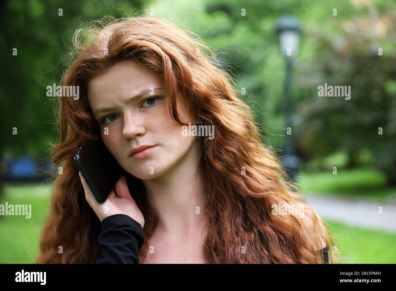 Frowning girl with long ginger hair and freckles talking on mobile phone in summer park looking into camera Stock Photo