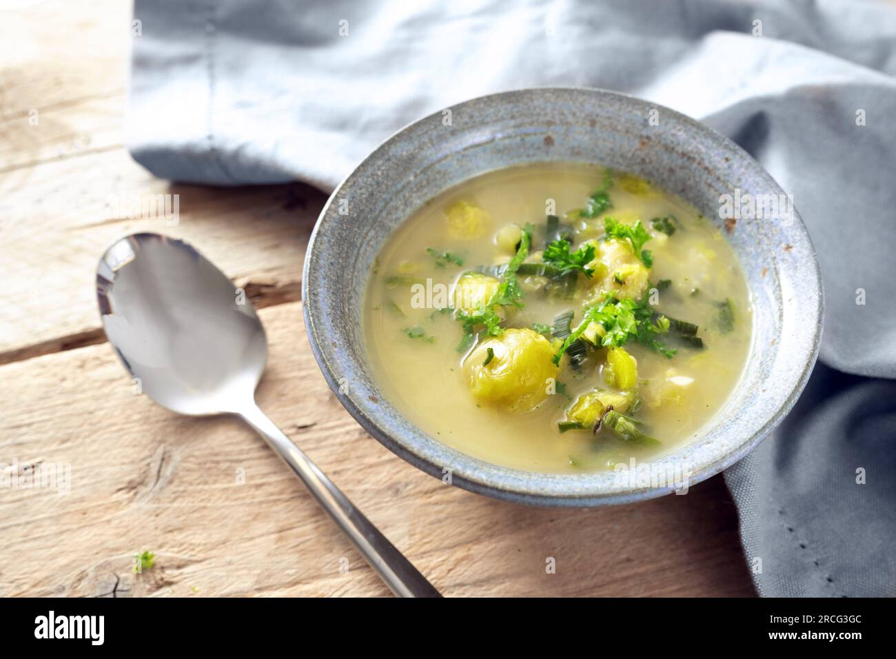 Vegetarian soup from vegetables like brussels sprout, leek and potato with parsley garnish in a blue bowl, spoon and napkin on a rustic wooden table, Stock Photo