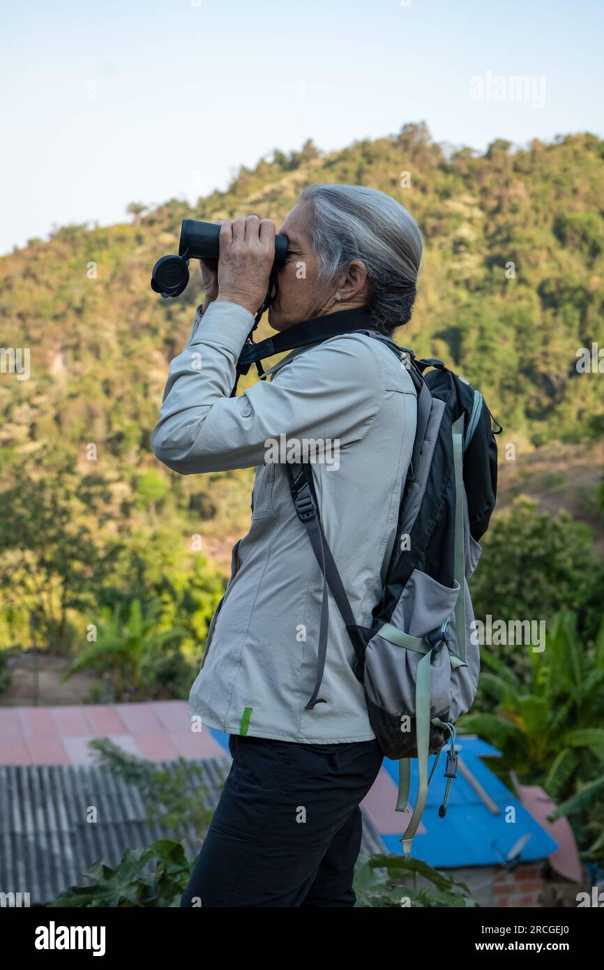 Elderly White Woman with Gray Hair Watches Birds Carefully with Binoculars by the Mountain Stock Photo