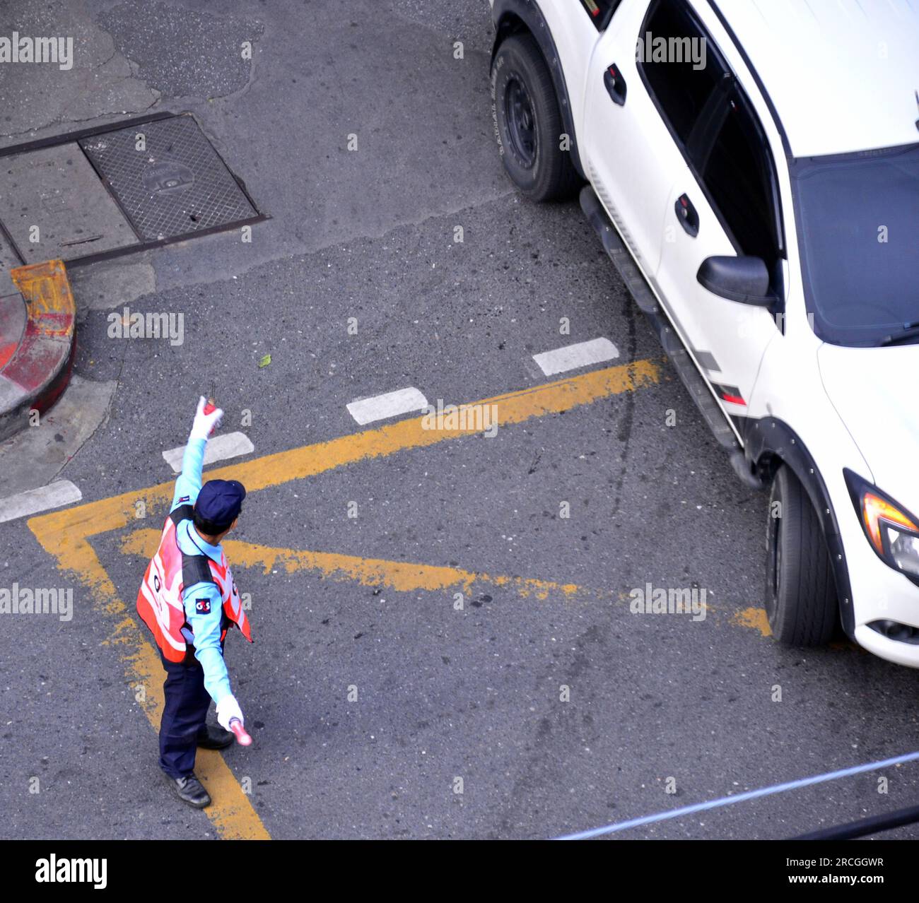 Overhead view of a male security guard, managing traffic on Surawong Road, Bangkok, Thailand, as a white car emerges from a side road to join the main road, named Surawong. Traffic accidents are common in Thailand and Surawong is a busy road, often filled with fast moving vehicles. Stock Photo