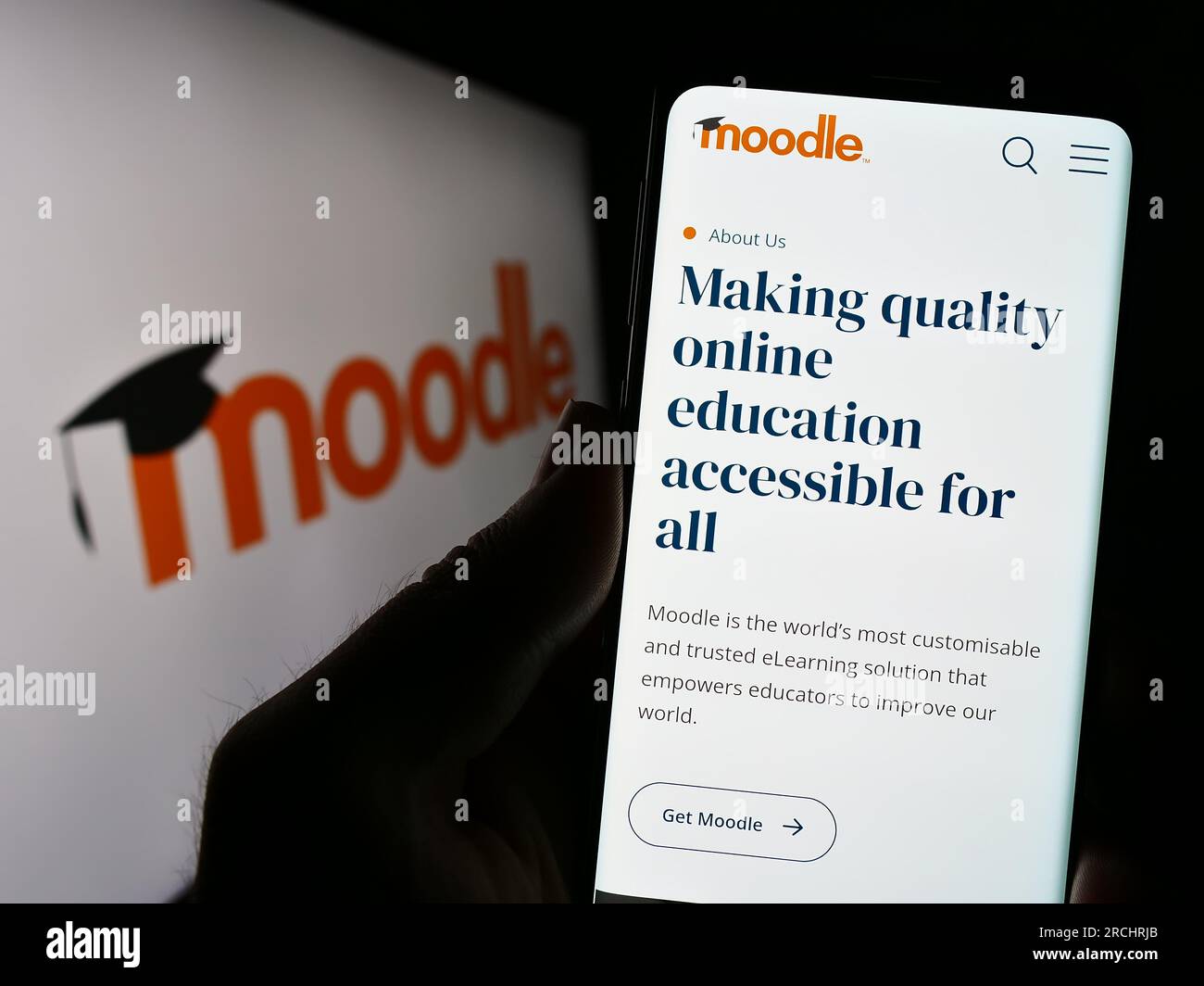 Person holding cellphone with webpage of learning platform Moodle on screen in front of logo. Focus on center of phone display. Stock Photo
