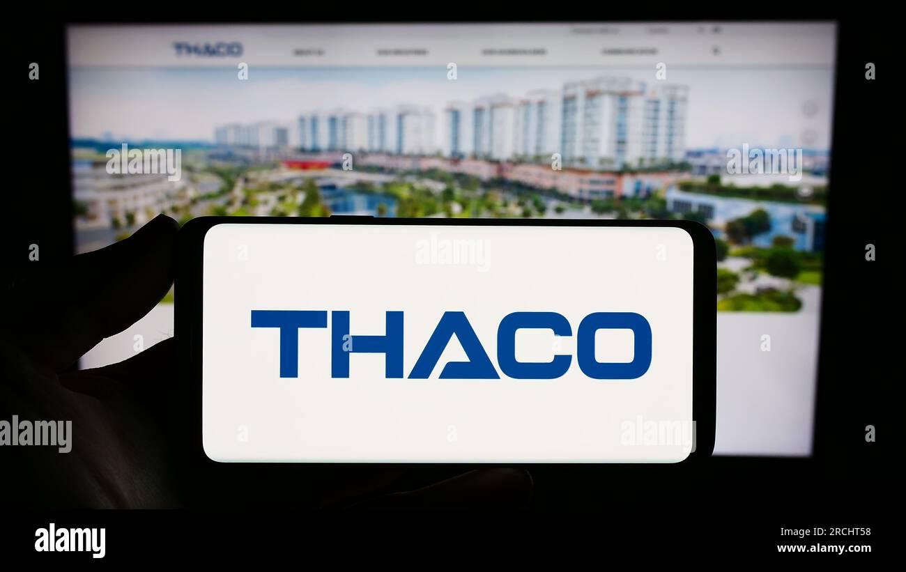 Person holding smartphone with logo of company Truong Hai Auto Corporation (THACO) on screen in front of website. Focus on phone display. Stock Photo