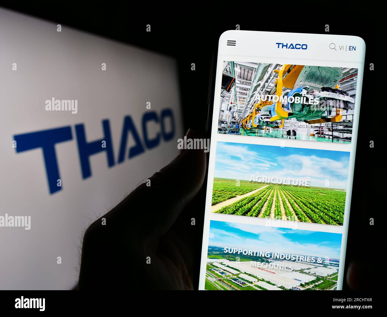 Person holding cellphone with website of company Truong Hai Auto Corporation (THACO) on screen in front of logo. Focus on center of phone display. Stock Photo