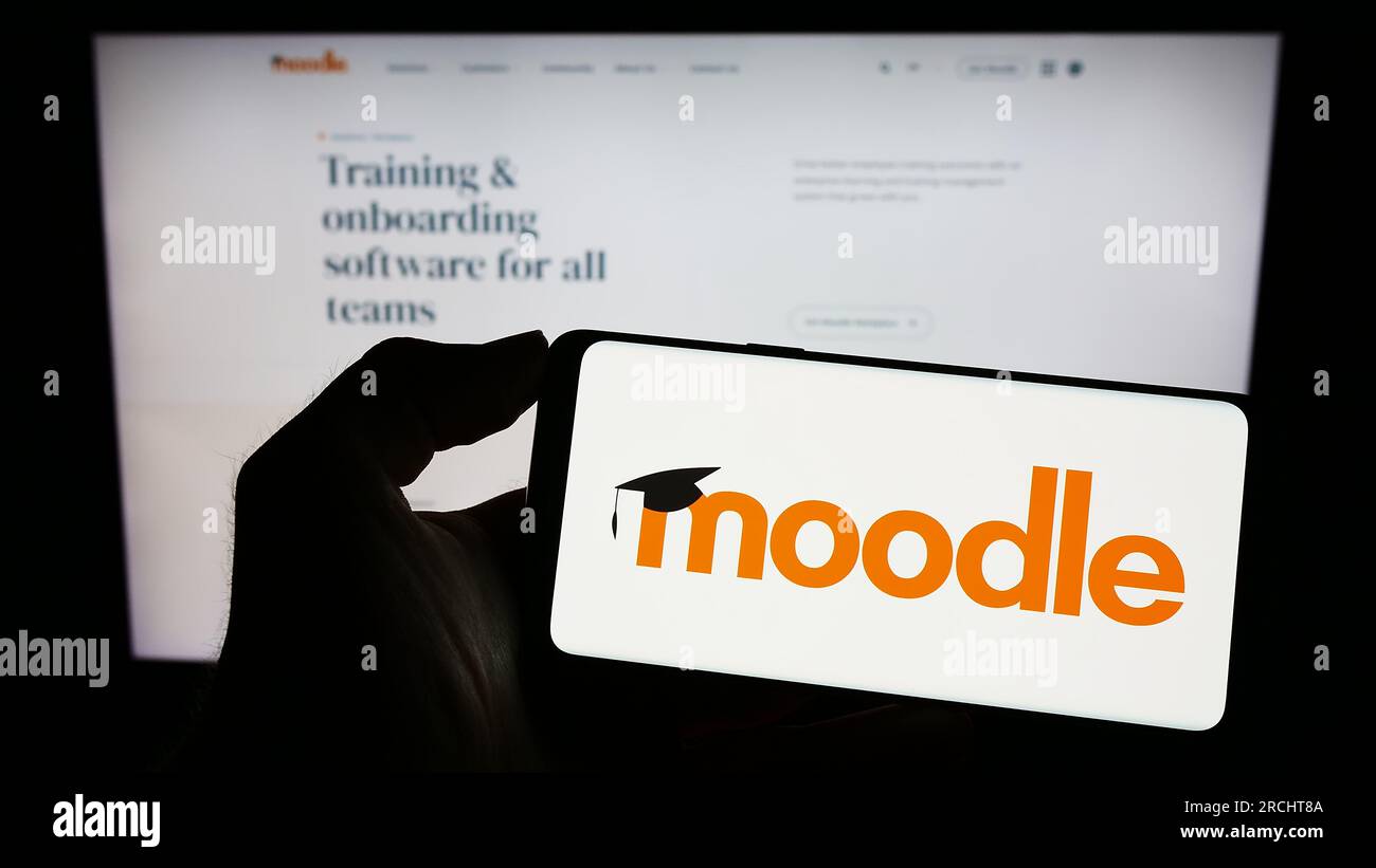 Person holding cellphone with logo of learning platform Moodle on screen in front of webpage. Focus on phone display. Stock Photo