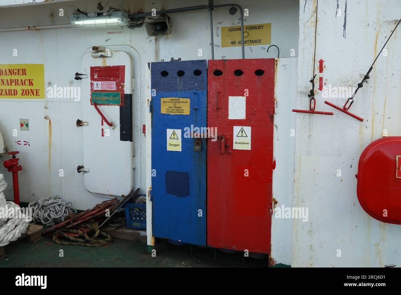 Stores of oxygen and acetylene gas bottles behind the blue and red metal doors with warning stickers situated on ship. Stock Photo
