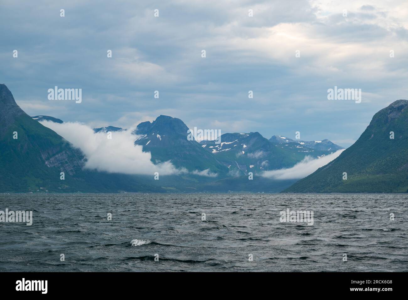 View from the sea towards Aldersund Norway with the island of Aldra to the right and mountains with fog and a cloudy sky in the background. Stock Photo