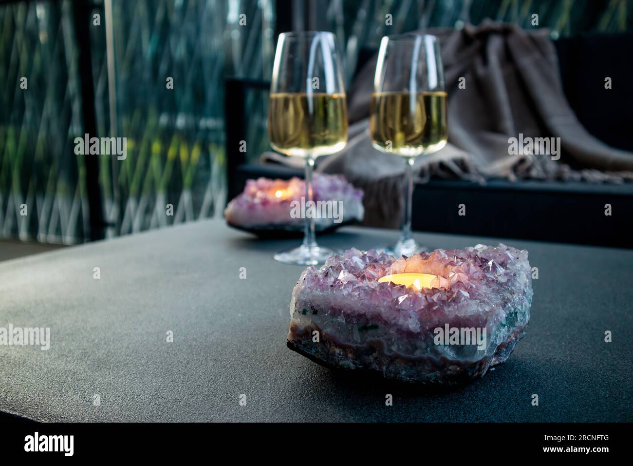 Selective focus on amethyst crystal geode candle holder with tea light candle burning inside, two white wine glasses on background in the evening. Stock Photo