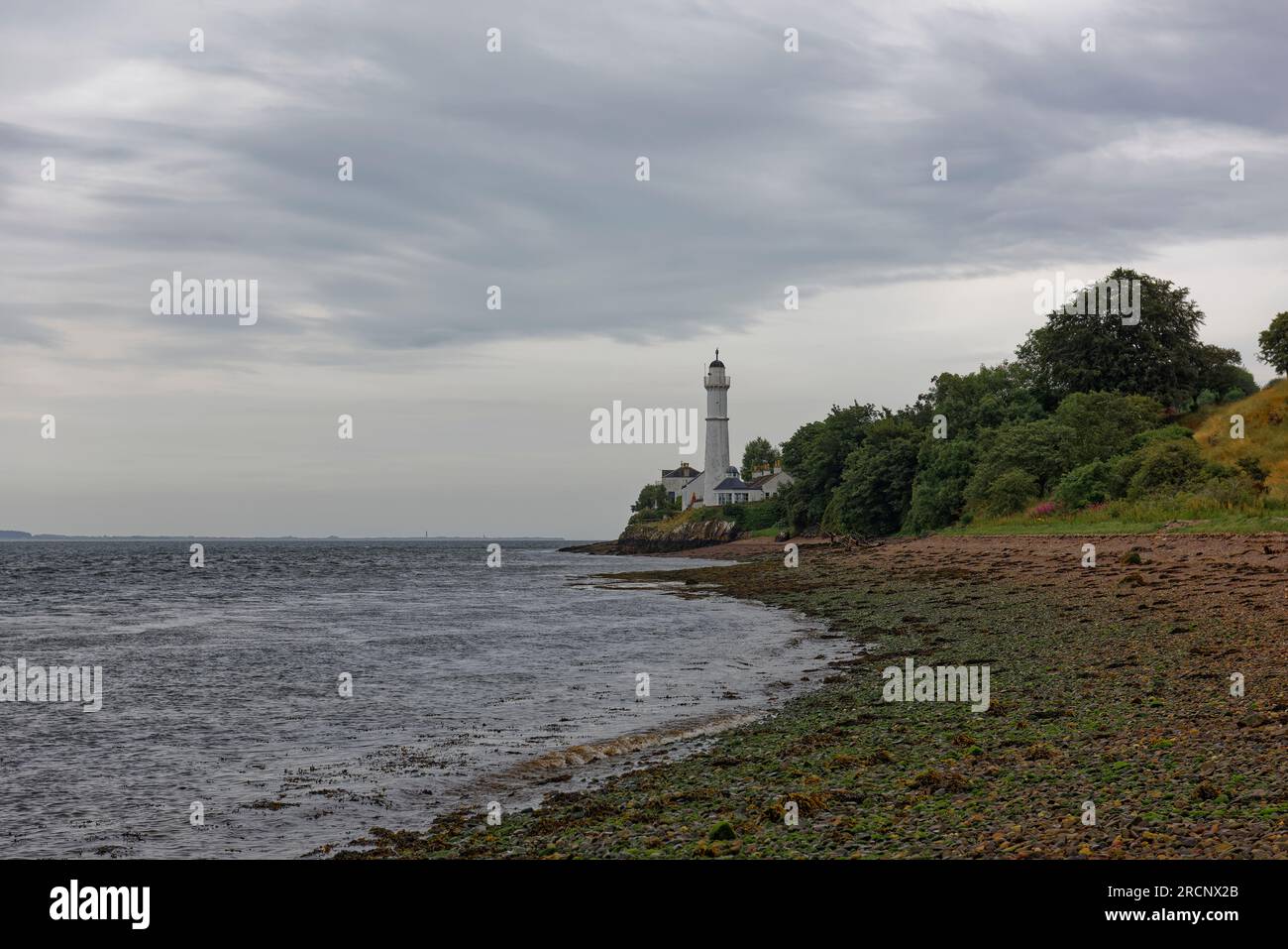 The West Lighthouse of the Tay Estuary on a Rocky outcrop above a curving shingle Beach littered with seaweed at low tide. Stock Photo