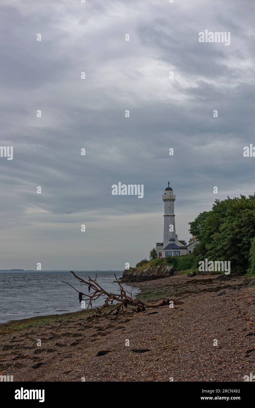 The West Lighthouse of the Tay Estuary on a Rocky outcrop above a curving shingle Beach with a Drift wood tree in the foreground. Stock Photo