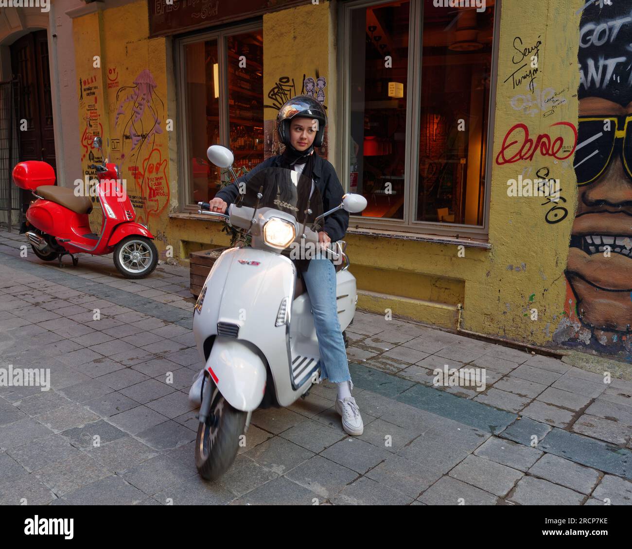 Young person on a white scooter with a red scooter behind on a street with a yellow wall in Istanbul, Turkey Stock Photo