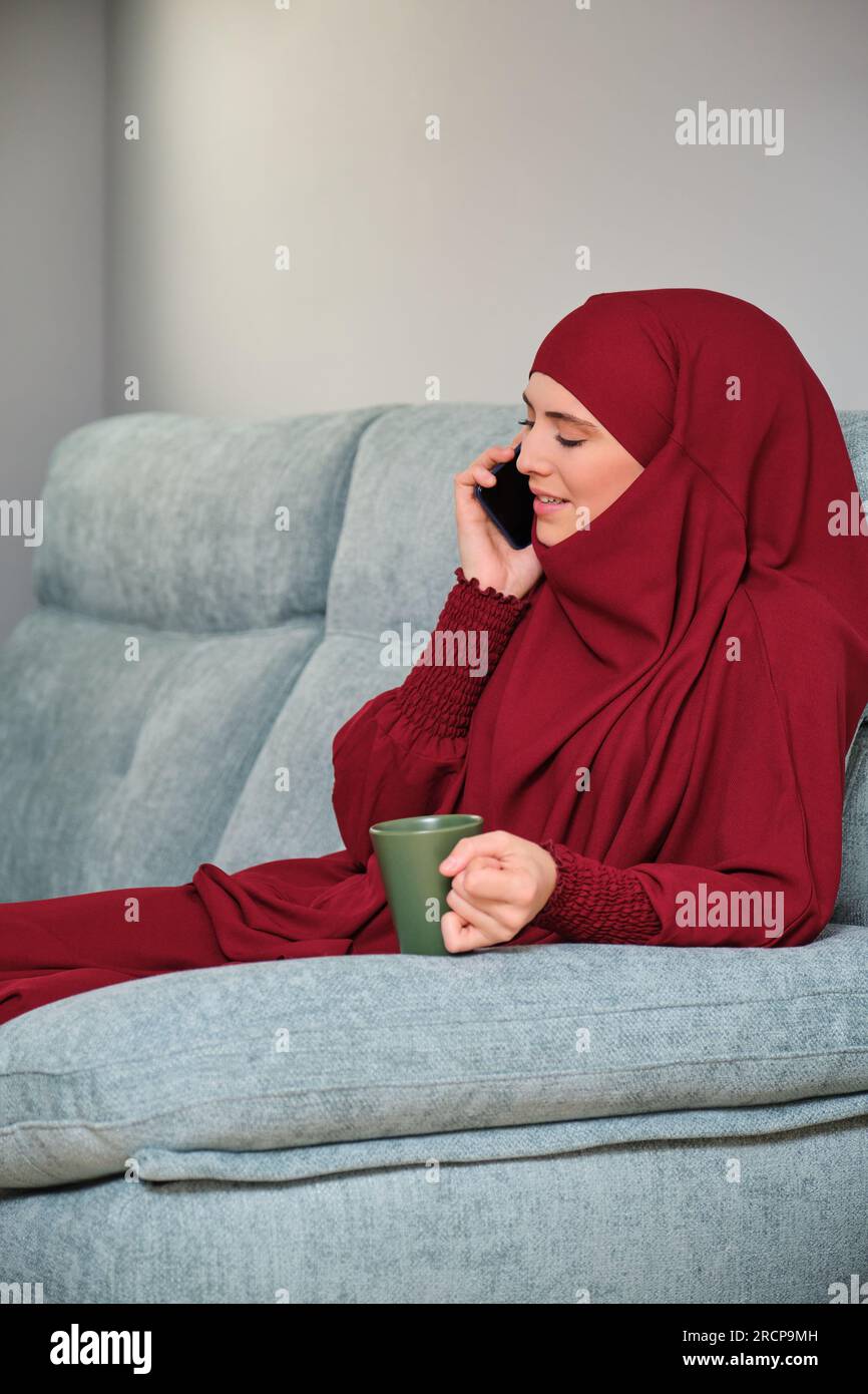 Muslim young woman in hijab speaking on the smartphone sitting on sofa ...