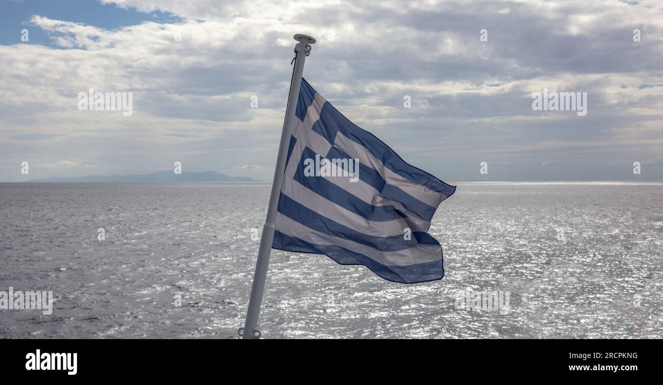 Greece sign symbol of leading shipping power in the world. Greek flag on flagpole waving over rippled sparkle sea water. Cloudy sky background. Stock Photo