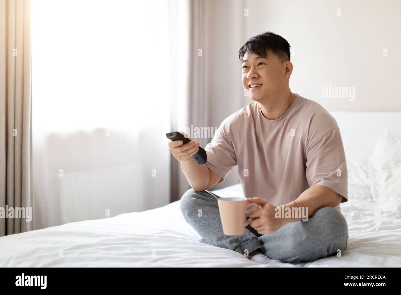 Happy middle aged asian man sitting on bed, watching TV Stock Photo