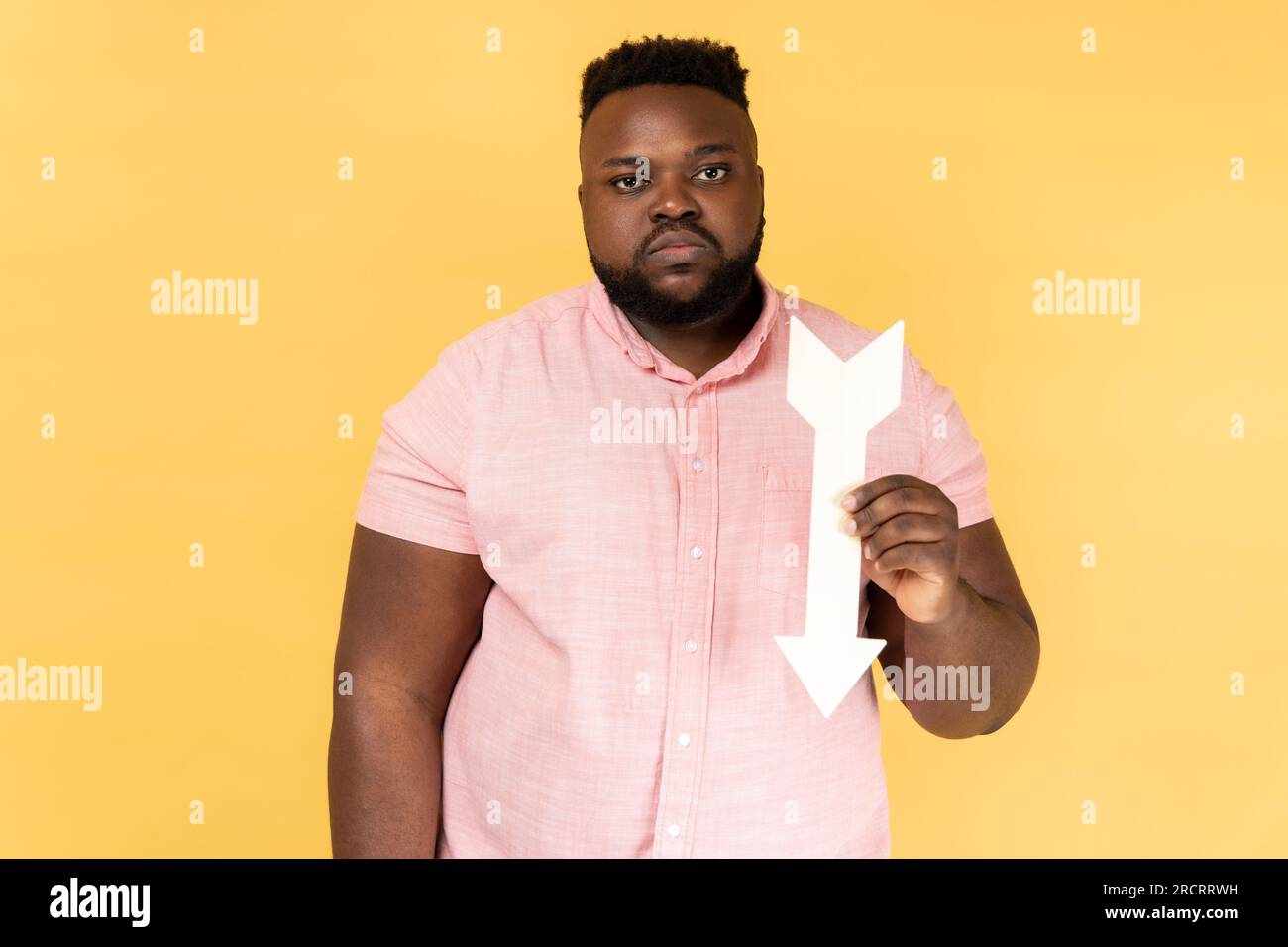 Portrait of sad upset depressed bearded man wearing pink shirt showing white arrow pointing down, expressing sorrow, downgrade concept. Indoor studio shot isolated on yellow background. Stock Photo