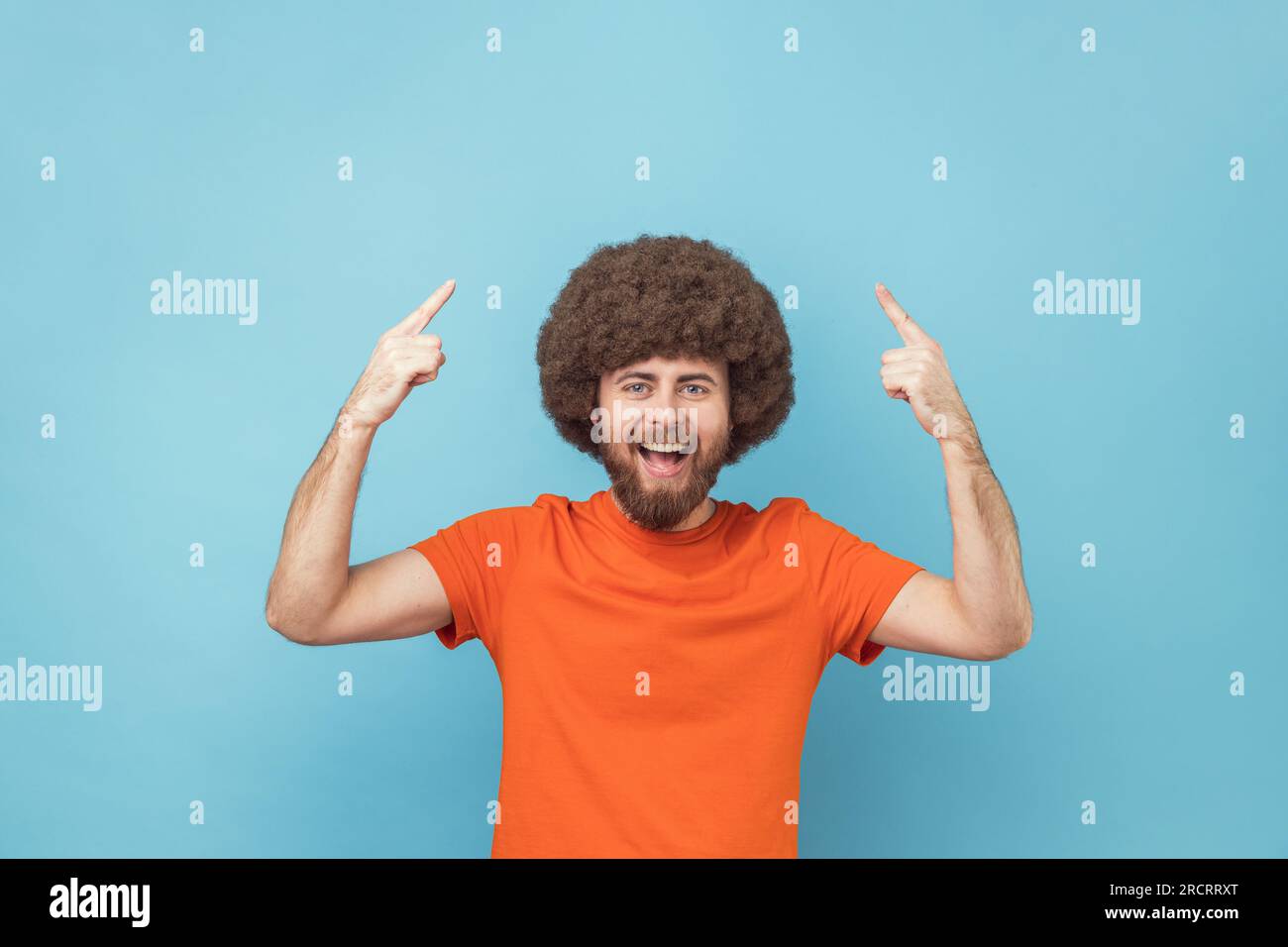 Portrait of extremely happy man with Afro hairstyle in orange T-shirt pointing finger up with toothy smile on face, clever smart man has idea, start up. Indoor studio shot isolated on blue background. Stock Photo