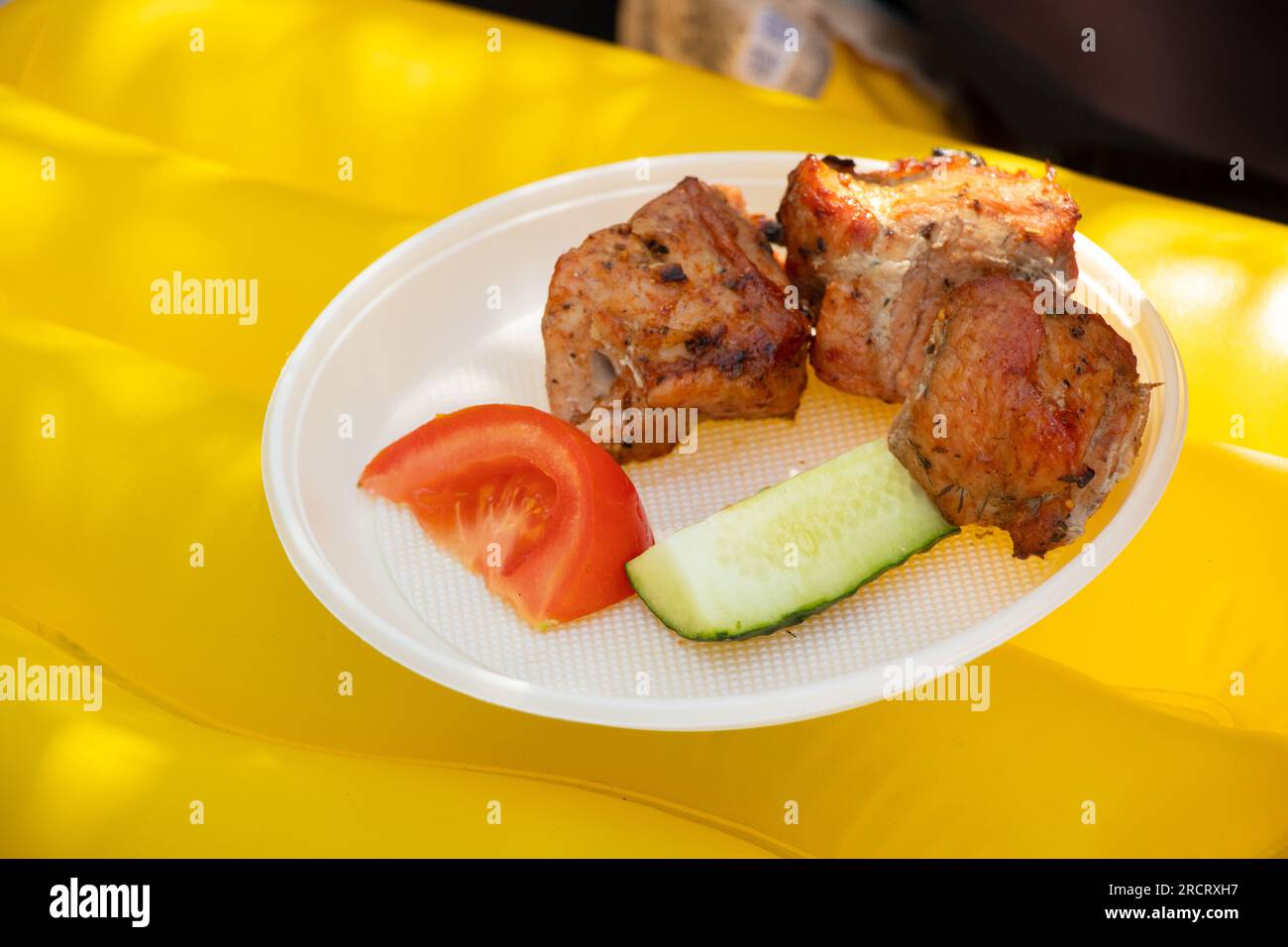 Grilled kebab and tomato with cucumber lie on a disposable plate on a yellow inflatable mattress in the summer at a picnic in Ukraine, food on the str Stock Photo