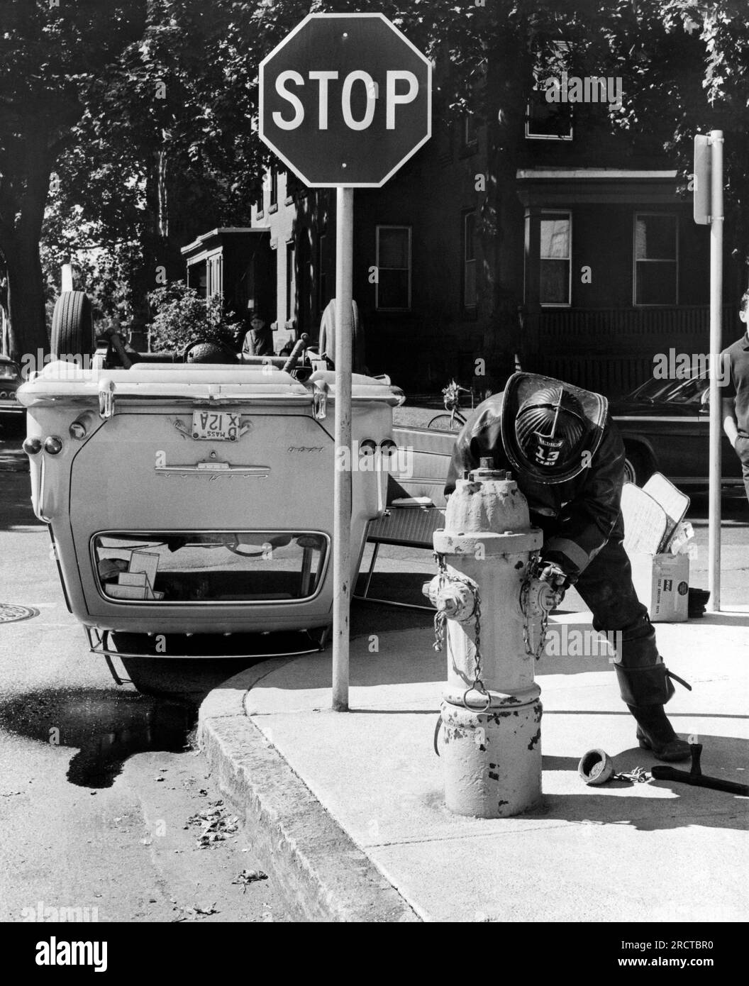 Worcester, Massachusetts:  October, 1967 An overturned car rests next to a stop sign, while a fireman prepares to wash away the spilled gasoline Stock Photo