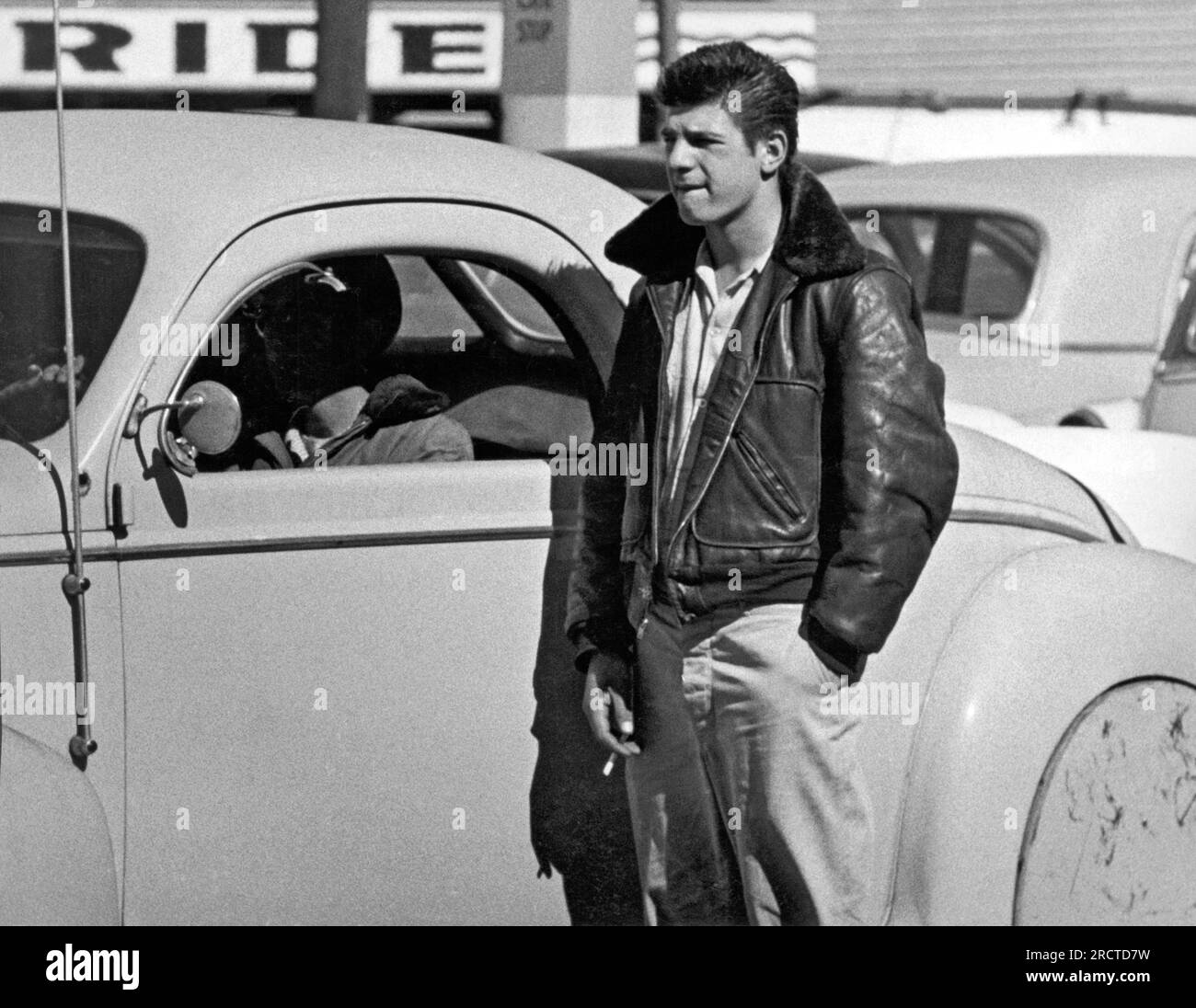 San Francisco, California:  c.1955 A young man with his hair slicked back and his leather jacket collar turned up standing next to a hot rod. Stock Photo