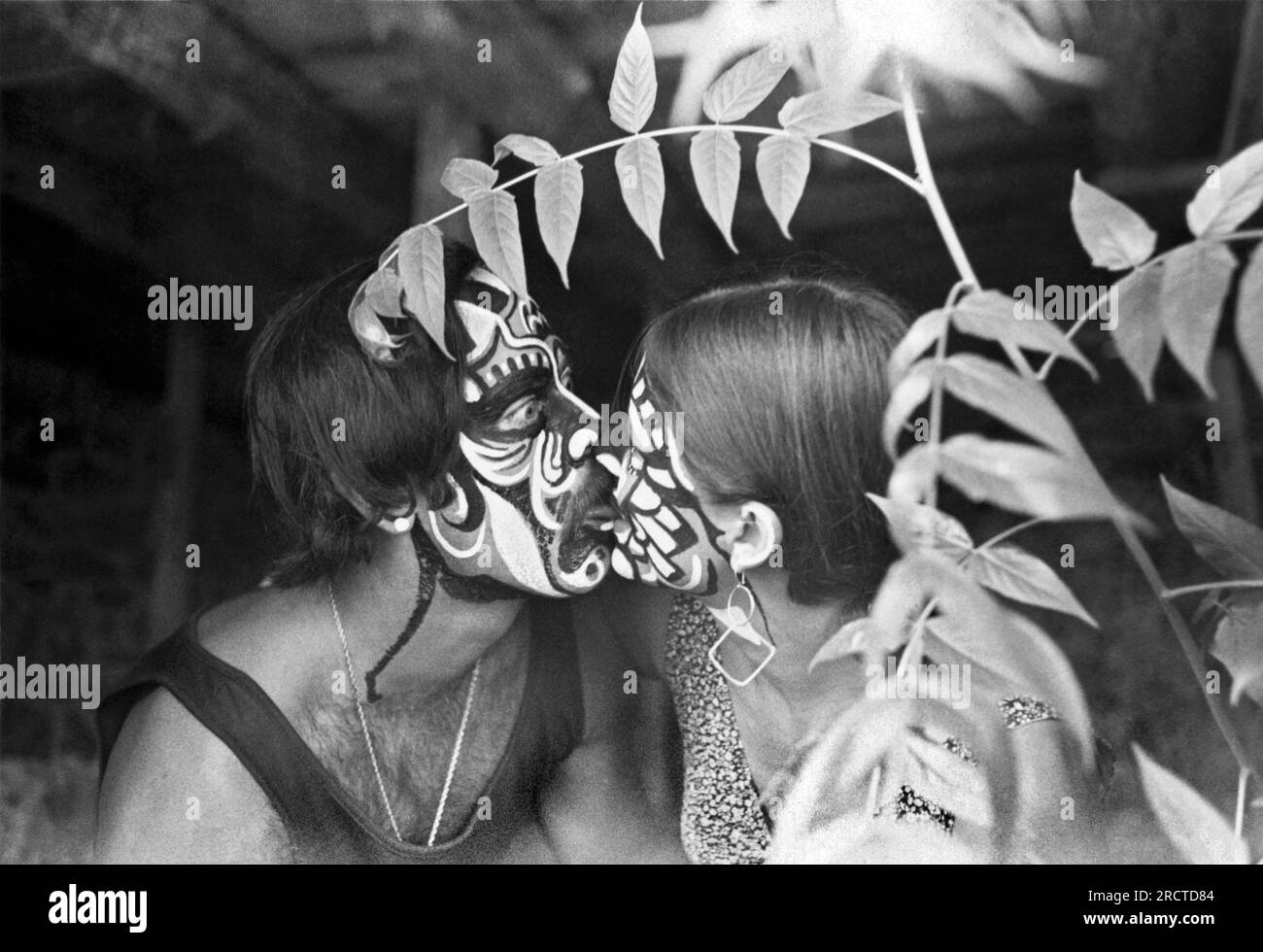 California:  c. 1969. A couple with painted faces touch tongues in the shrubbery. Stock Photo