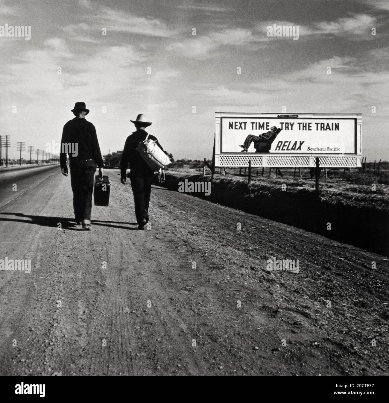 California:  March, 1937 Two men, each carrying luggage, walk along a highway towards Los Angeles while a Southern Pacific railroad billboard shows a man reclining comfortably in a railroad passenger car seat and states, 'Next Time Try the Train, Relax'. Stock Photo