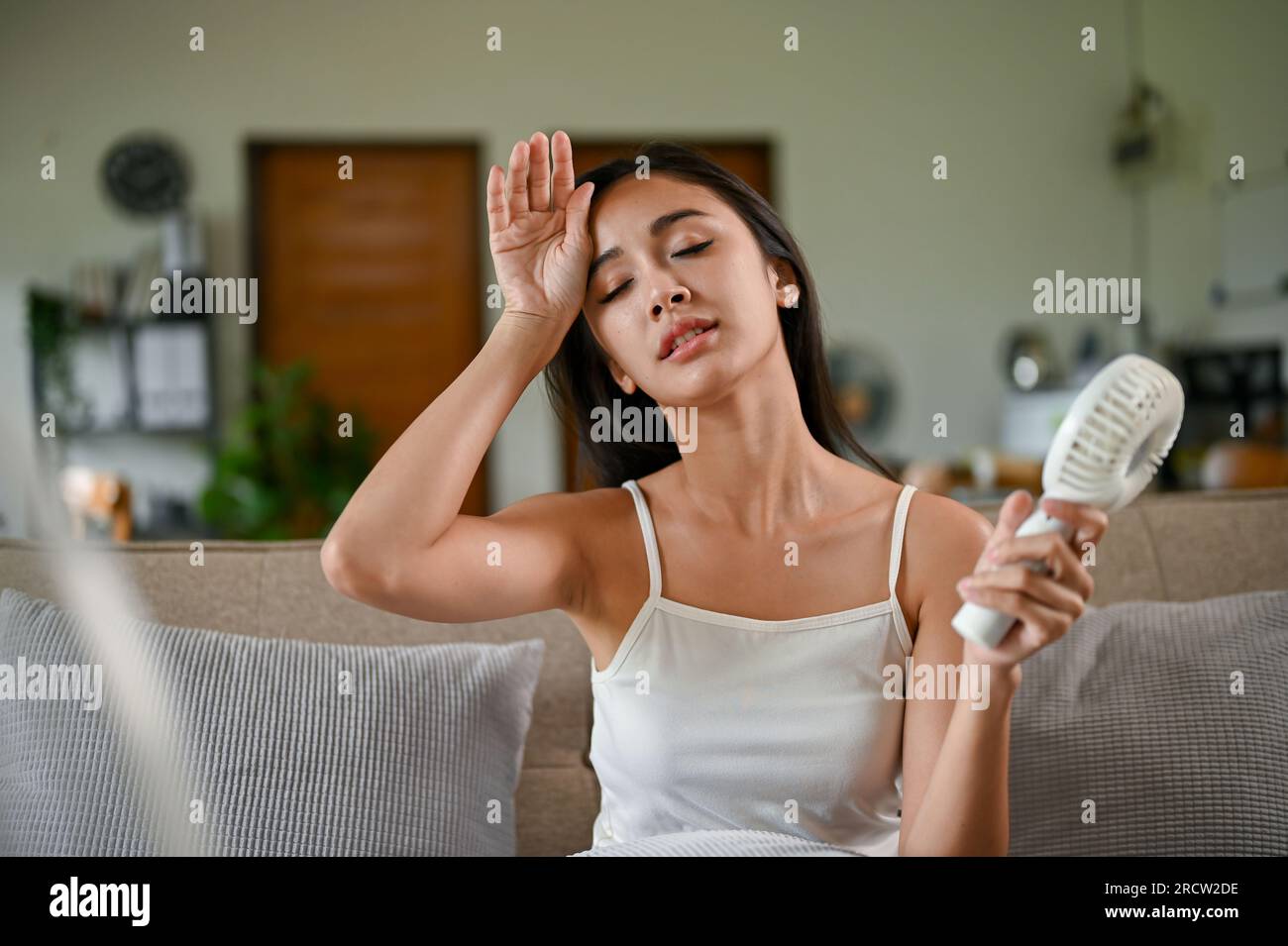 A tired and overheated Asian woman suffering from a heat attack uses an electric handy fan to cool herself down while resting on a couch in her living Stock Photo