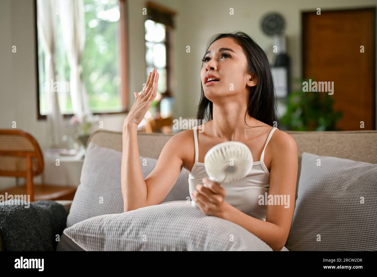 An overheated Asian woman suffering from a heat attack uses an electric handy fan to cool herself down while resting on a couch in her living room on Stock Photo