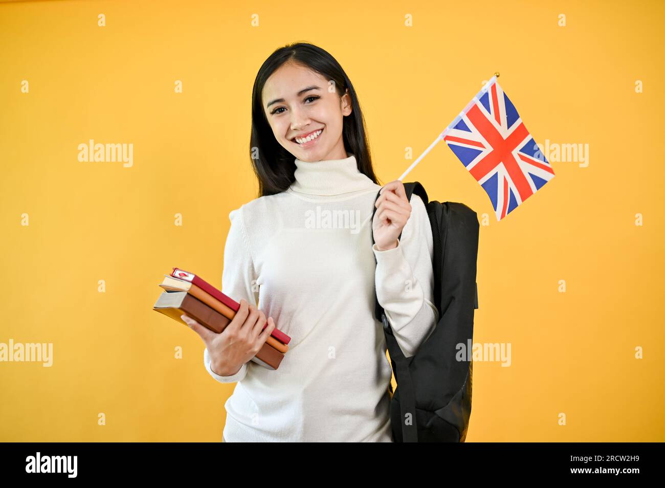 A pretty Asian female college student is holding her books and the United Kingdom flag, standing against a yellow, isolated background. Exchange stude Stock Photo