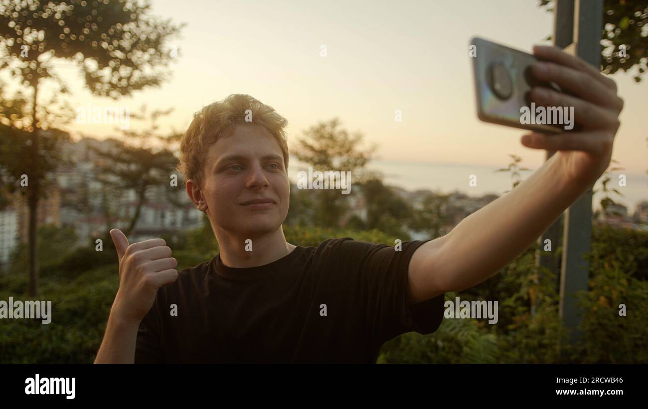 Capturing Moments: Handsome Blond Young Man Taking Selfies with His Smartphone, Striking Various Poses Outdoors Stock Photo