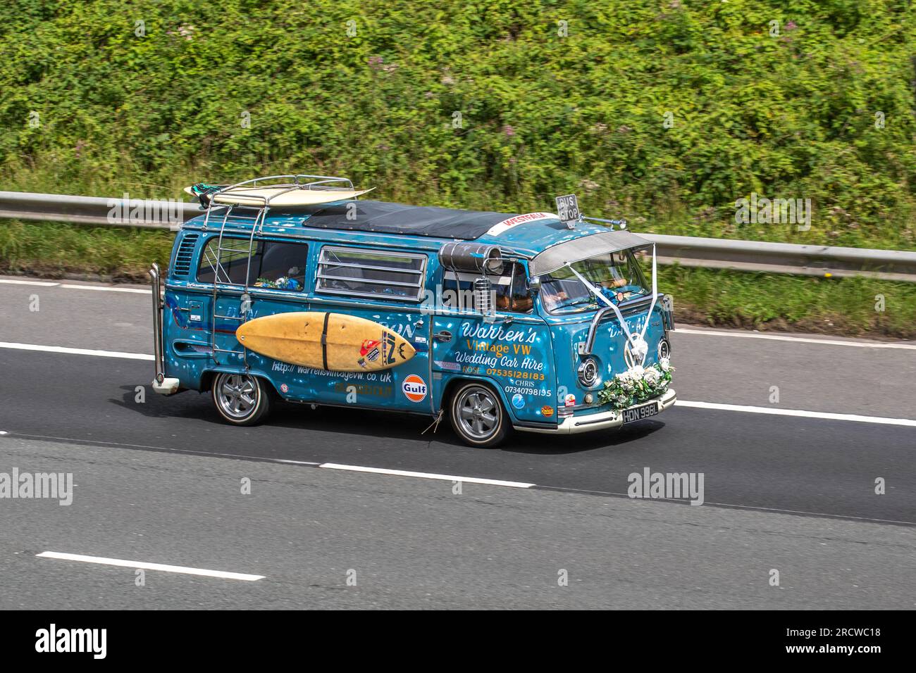 Warren's Vintage VW. 1973 70s seventies Blue Custom VW Volkswagen Motor Caravan LCV Petrol 1584 cc. A curious wedding hire car with hippie decorations, white ribbons & surfboard; travelling at speed on the M6 motorway in Greater Manchester, UK Stock Photo