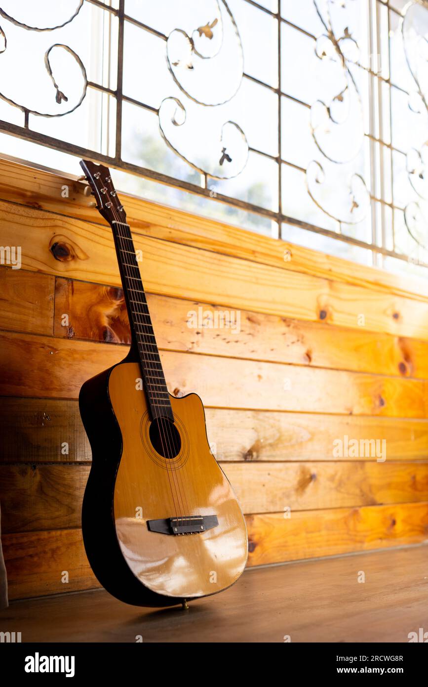 Acoustic guitar leaning against wood panelled wall below window in sunny cabin, copy space Stock Photo