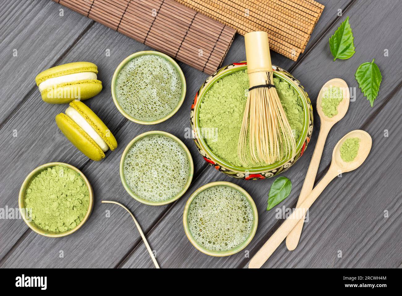 Bamboo whisk in bowl with matcha powder. Matcha green tea, macaroni cakes and wooden spoons on table. Flat lay. Dark wooden background Stock Photo