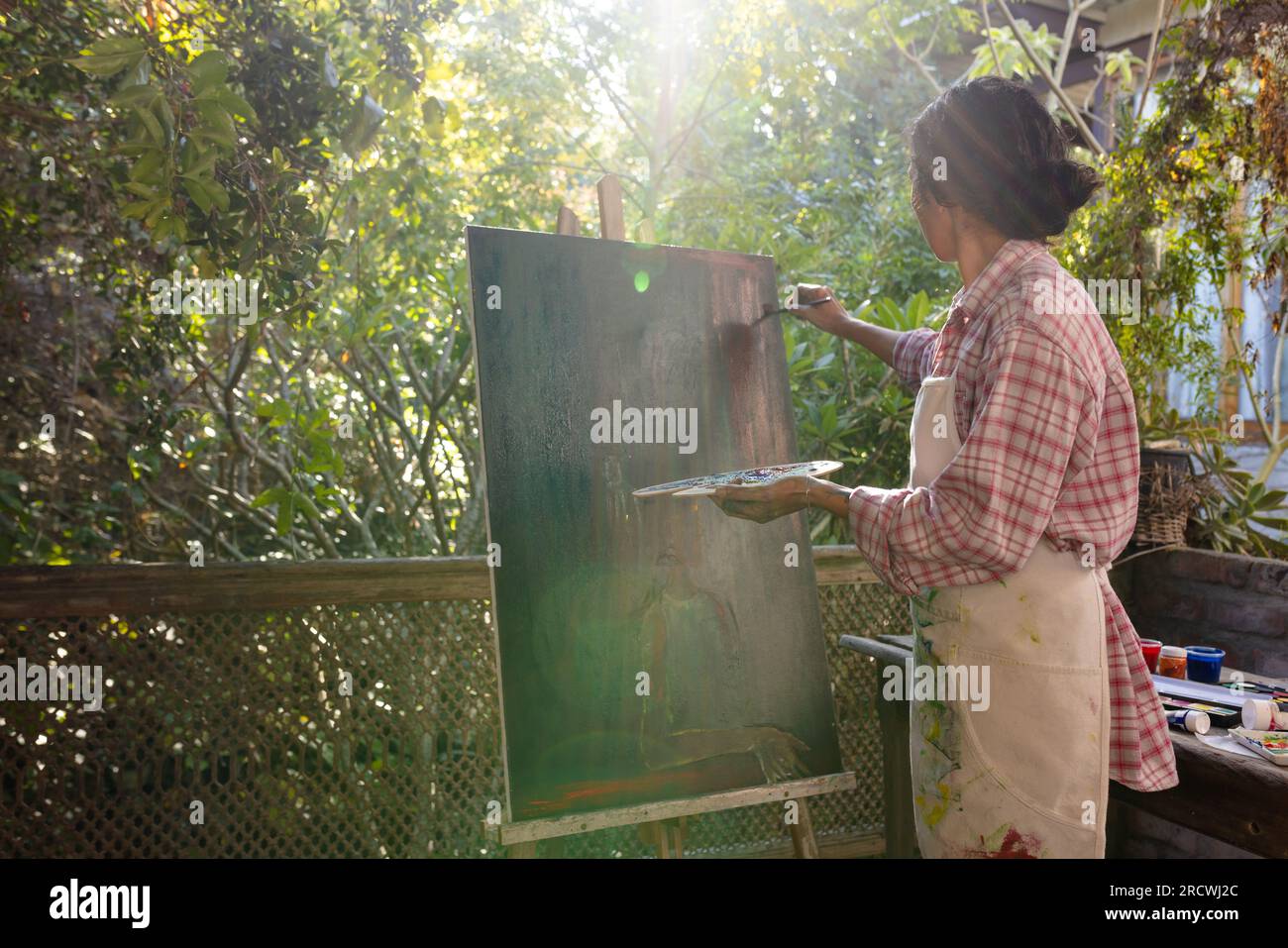 Biracial woman wearing apron painting on easel using brush and colour