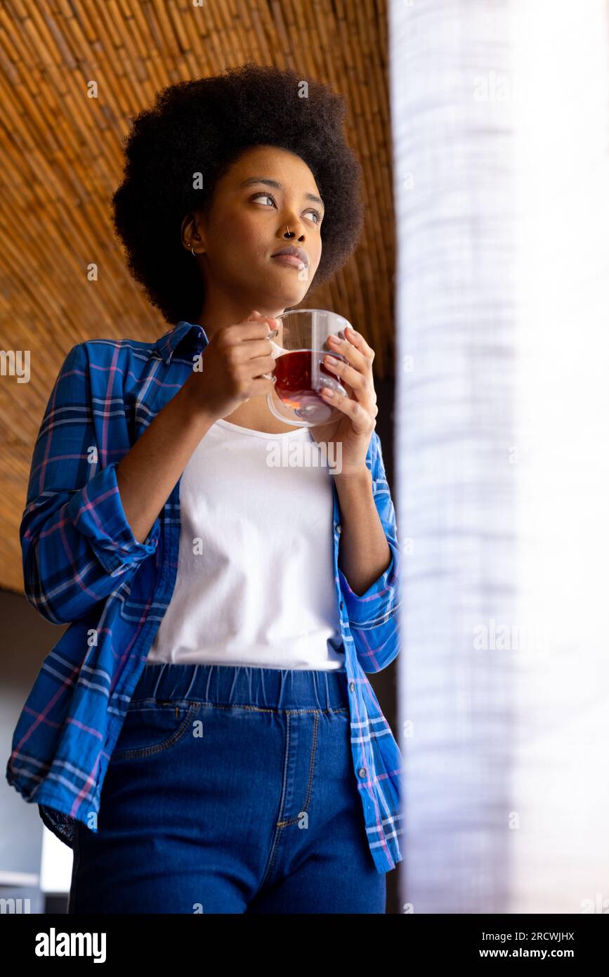 Thoughtful african american woman standing with cup of tea, looking out of window Stock Photo