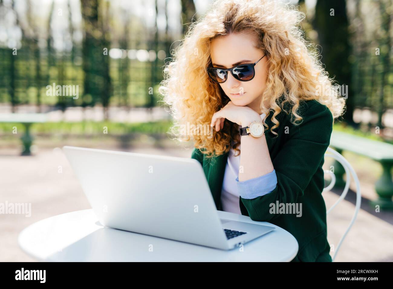 Curly-haired woman in green jacket, sunglasses, reading news on laptop outdoors. Tech-savvy, contemplative. People, technology. Stock Photo