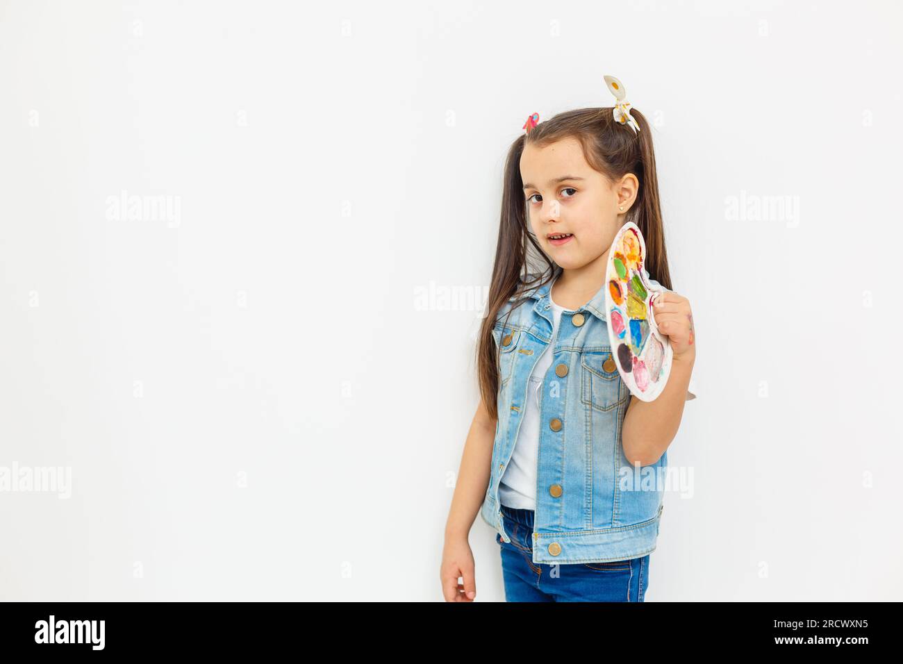 A joyful little girl with a paint palette and a brush on a white ...