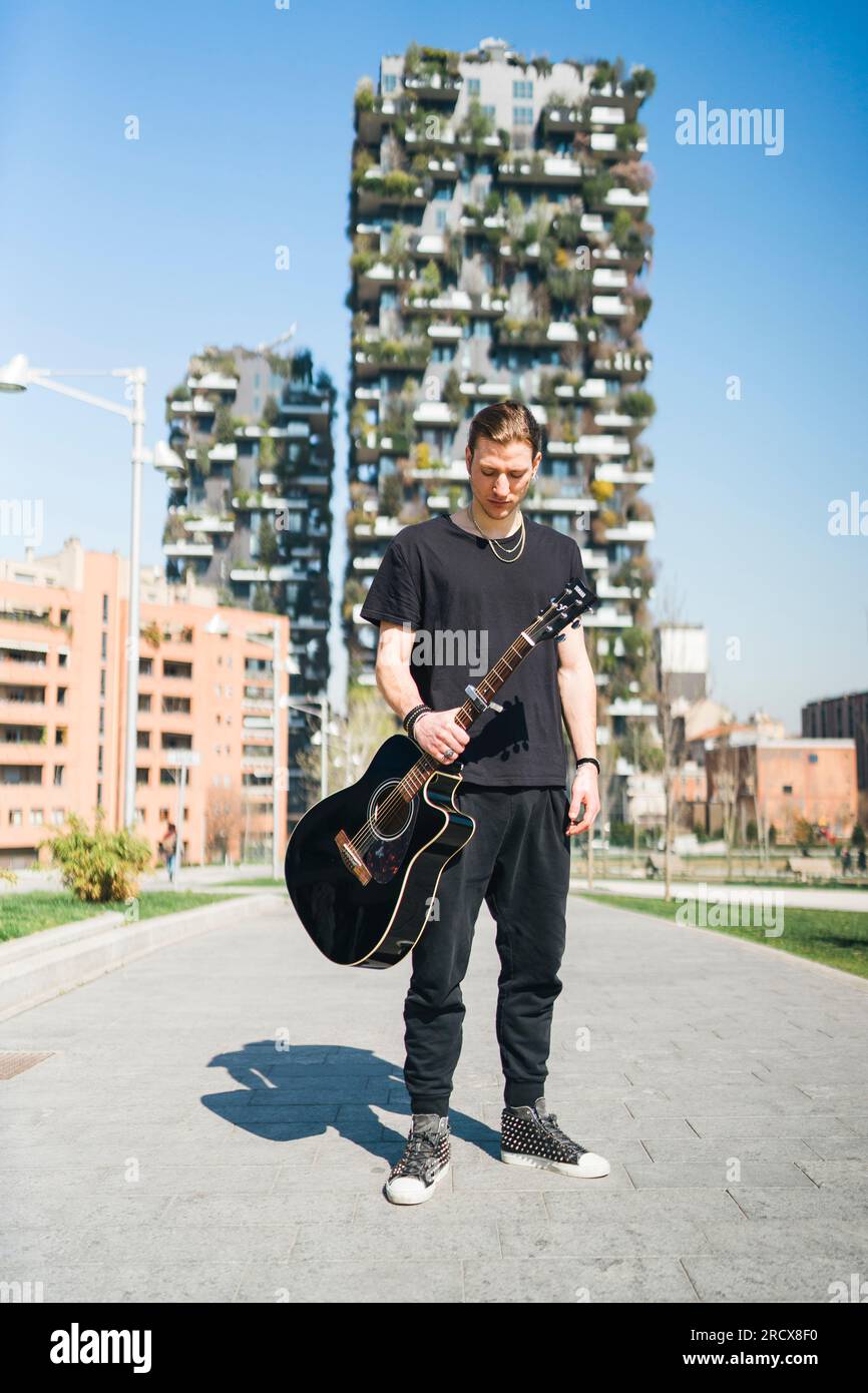 man holding a guitar is standing in front of two green skyscreapers Stock Photo