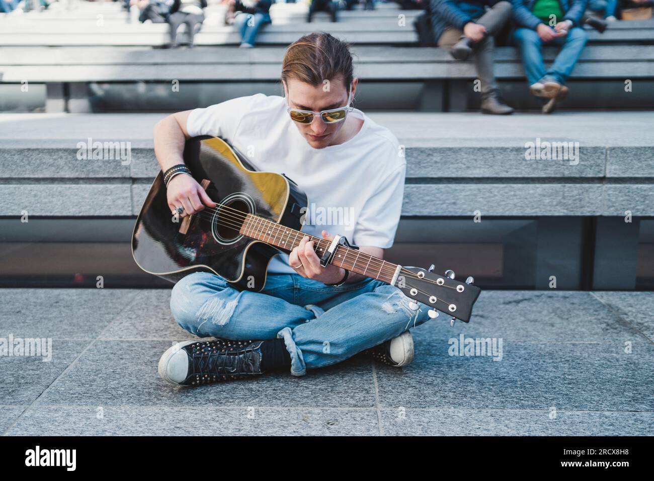 musician looking at the guitar sitting on the floor with people behind Stock Photo