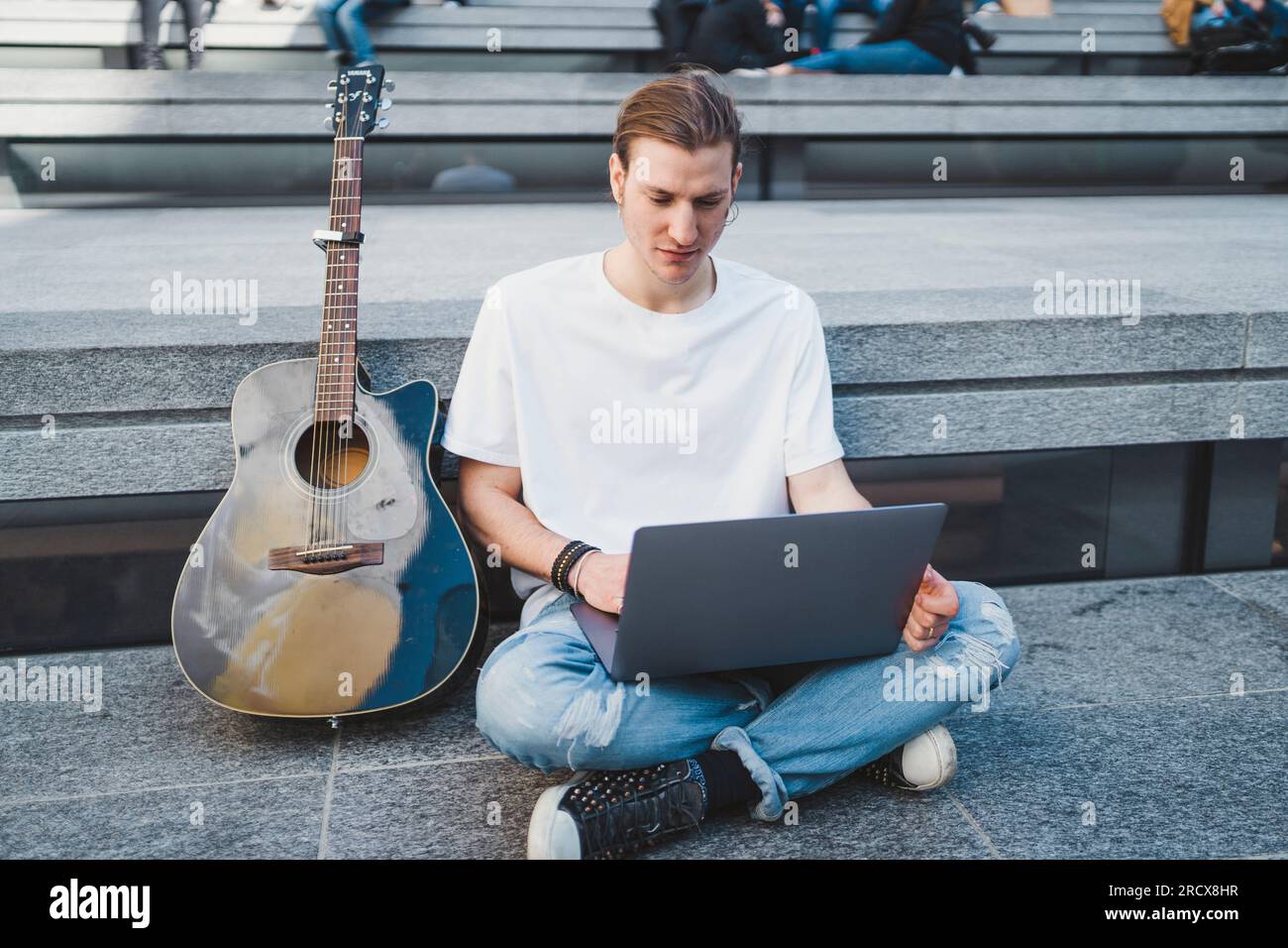 white man using laptop computer in square beside his classical guitar Stock Photo
