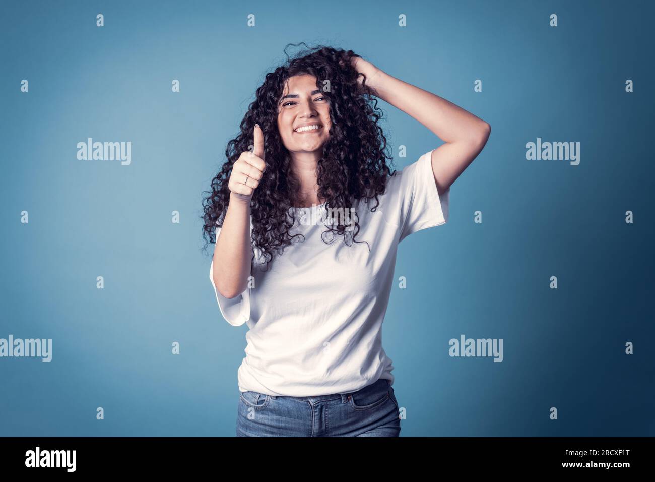 Shot of happy confident curly woman with toothy smile, wears casual basic solid white t-shirt, expresses good emotions, enjoys nice day, doing thumb u Stock Photo