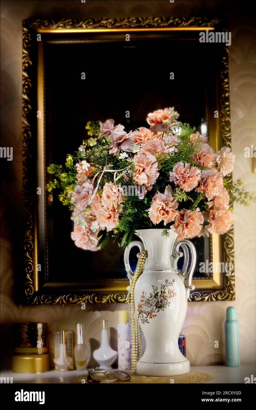 Artificial flowers in a powder pink color stand in a tall vase in front of a mirror and on a chest of drawers. Retro decor, rustic. Stock Photo
