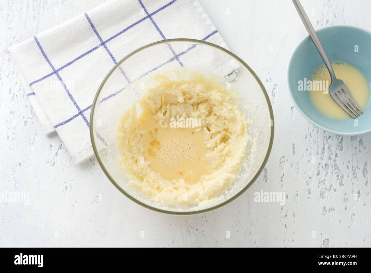 Glass bowl with whipped butter with sugar and beaten egg on a light blue background, top view. Cooking homemade milk cakes, do it yourself, step by st Stock Photo