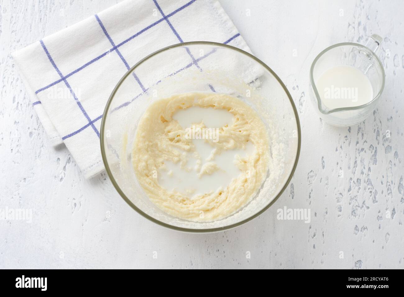 Glass bowl with whipped egg-cream-sugar mass, adding milk on a light blue background, top view. Cooking homemade milk cakes, do it yourself, step by s Stock Photo