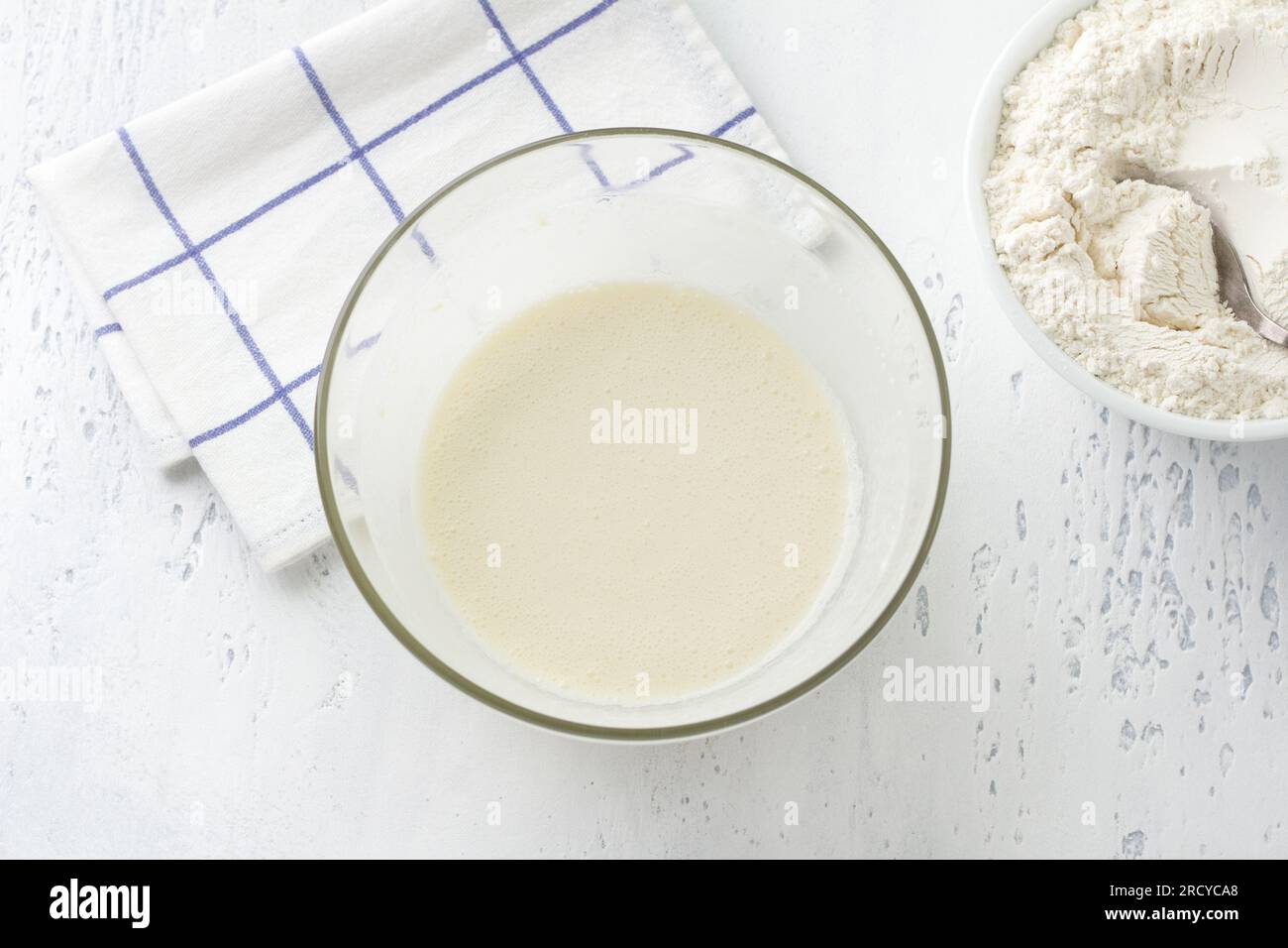A glass bowl with a whipped mass of eggs, butter, sugar and milk on a light blue background, top view. Cooking homemade milk cakes, do it yourself, st Stock Photo