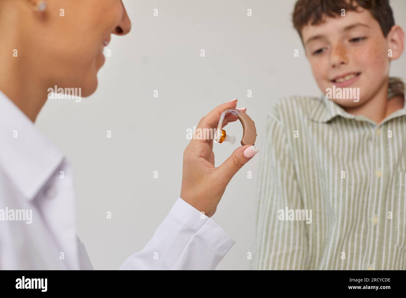 Modern effective hearing aid in hands of female ENT doctor showing it to smiling preteen boy. Stock Photo