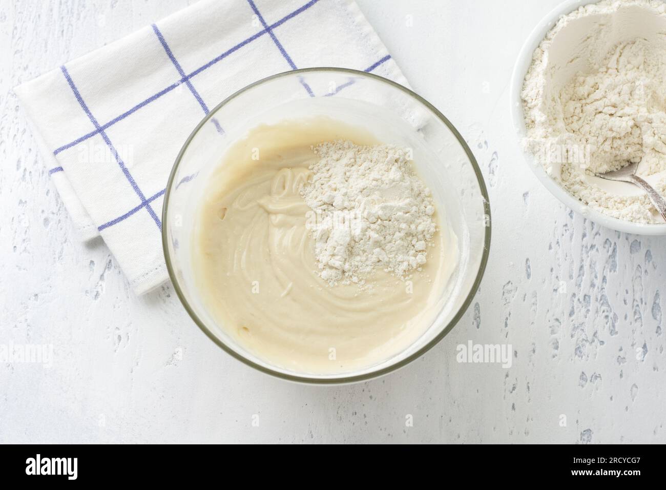 Glass bowl with dough, adding flour on a light blue background, top view. Cooking homemade milk cakes, do it yourself, step by step, step 6. Stock Photo