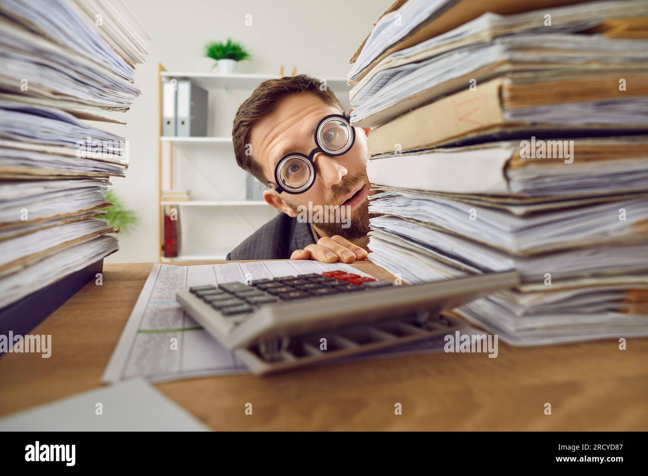 Funny shy male bookkeeper in glasses hiding behind piles of paperwork on his office desk Stock Photo