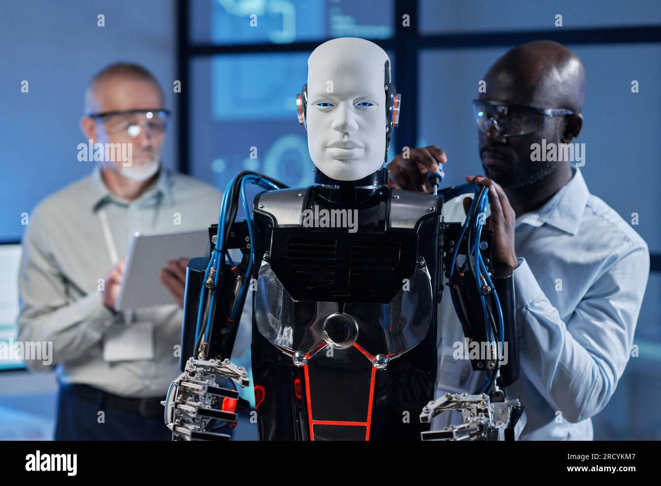 African American engineer repairing robot with tools while his colleague programming it in background Stock Photo