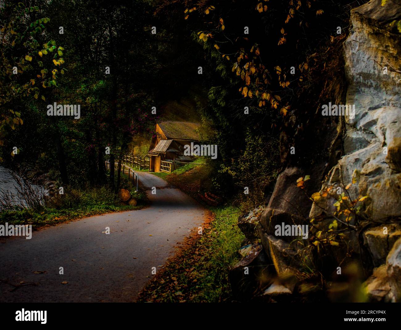 A lonely road next to rocks and a small wooden house. Far in the mountains in the countryside. A fabulous place in the shade of trees and mountains. Stock Photo