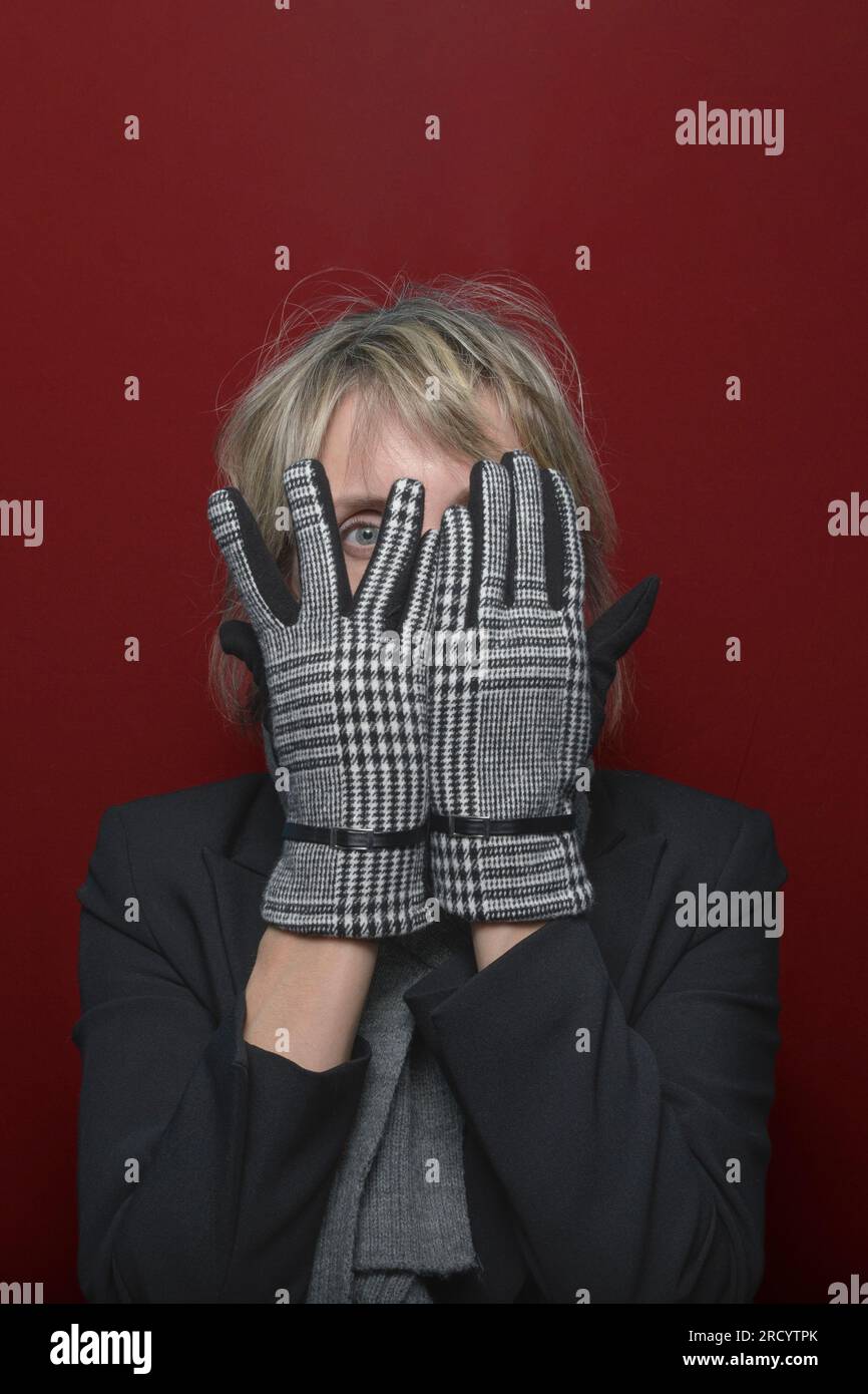 Turn a blind eye. A female model in elegant gloves looks through her fingers at the camera. portrait on a red background Stock Photo